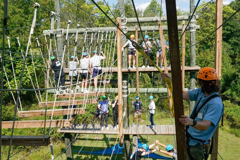 The image shows an outdoor adventure park with people participating in a ropes course. Participants are navigating various obstacles, including rope bridges and platforms, suspended high above the ground. The course is set amidst a lush green forest, with trees visible in the background. People are wearing helmets and safety harnesses, indicating safety precautions.